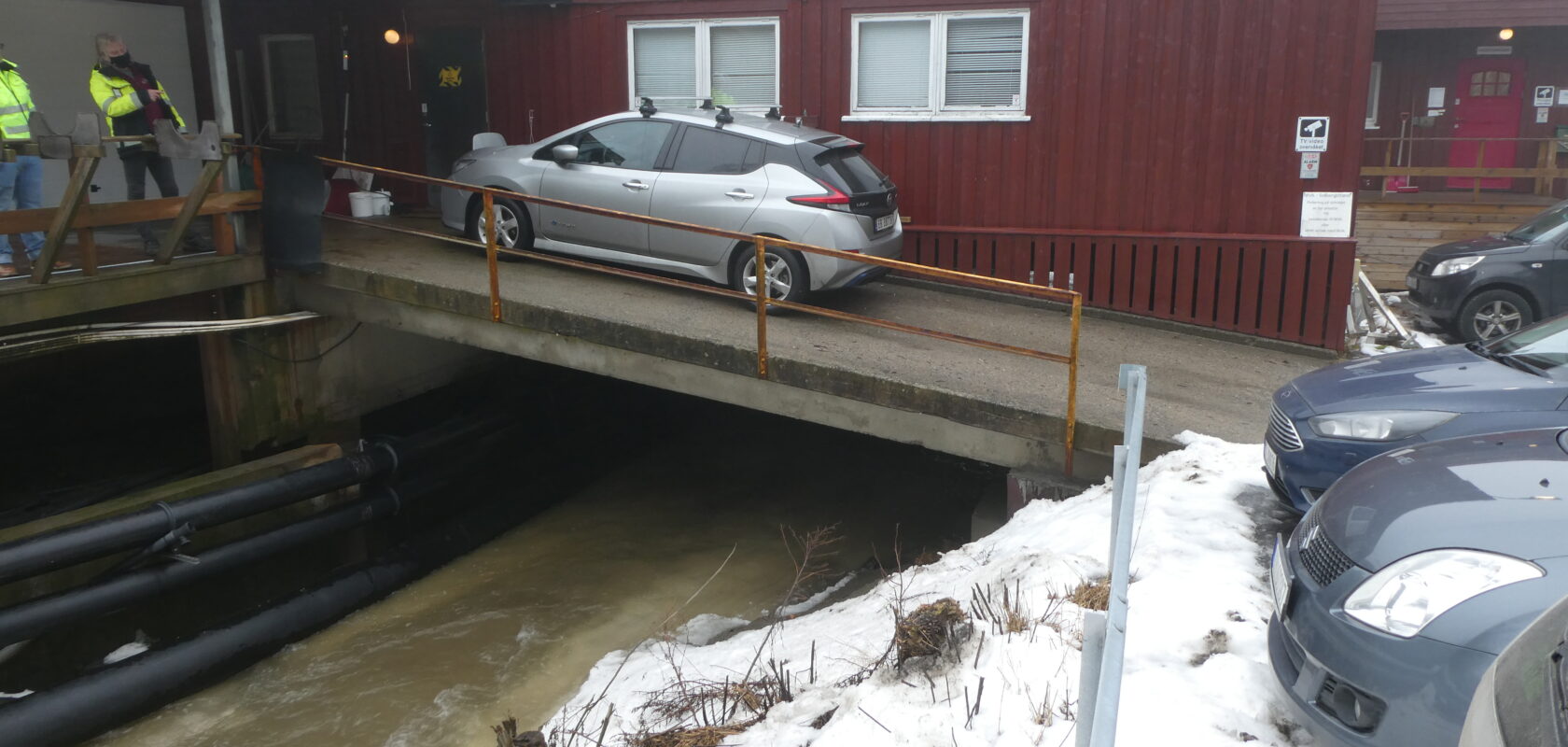 Bildet viser utsiden av forskningstasjonen. Det står en bil parkert på en bru som går opp til det røde bygget. Under renner det vann.